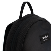 Close-up of a black Flowfold backpack showing the top handle, zipper, and a black brand patch with white text on a white background.