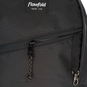 Close-up of a black Flowfold backpack showing a front zippered pocket with a black zipper pull, a silver-tone carabiner, and the Flowfold brand patch on the upper left.