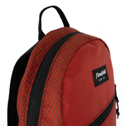 A close-up of a red and black Flowfold backpack, showing a red and black checkered pattern panel, black zippers, and a black Flowfold brand patch on a white background.