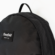 Close-up of a black Flowfold backpack showing the top handle, textured fabric, and a black brand patch with white text reading 'Flowfold MAINE USA' on a white background.