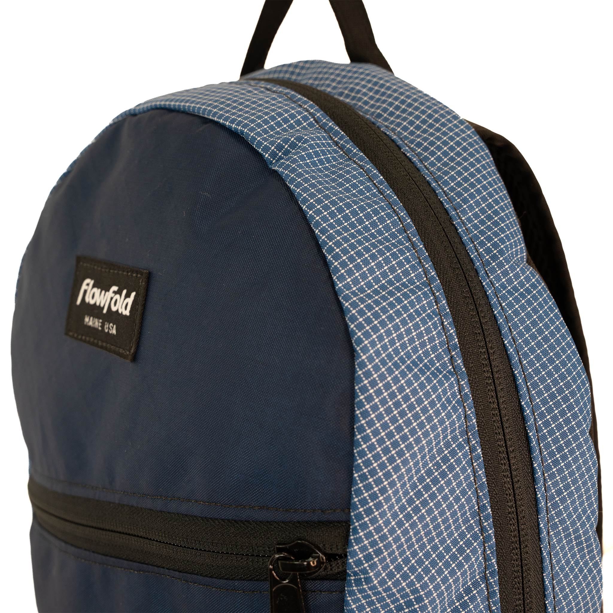 Close-up of a Flowfold backpack showing a navy blue front panel, a blue-and-white grid patterned accents, black zippers, and a black Flowfold brand patch.